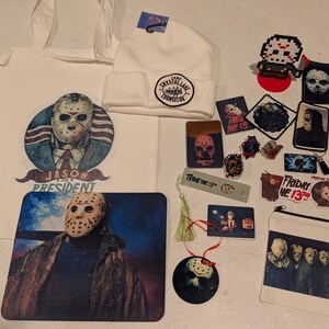 May include: A collection of Friday the 13th themed merchandise including a white tote bag with a Jason Voorhees graphic, a white beanie with a Camp Crystal Lake patch, a mousepad, various stickers, patches, a bookmark, and a zippered pouch.