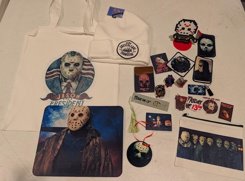 May include: A collection of Friday the 13th themed merchandise including a white tote bag with a Jason Voorhees graphic, a white beanie with a Camp Crystal Lake patch, a mousepad, various stickers, patches, a bookmark, and a zippered pouch.