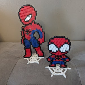 Puede incluir: Dos figuras de Spiderman rojas y azules hechas con cuentas de perler, de pie sobre telara&ntilde;as de cuentas de perler blancas.