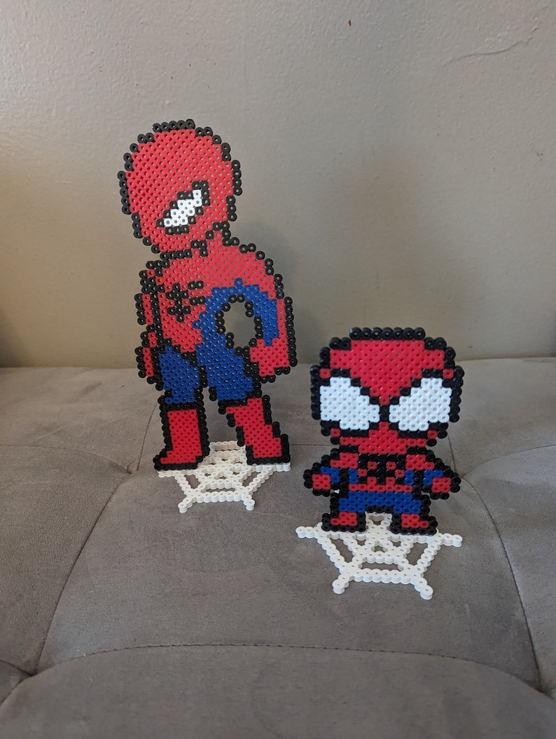 Puede incluir: Dos figuras de Spiderman rojas y azules hechas con cuentas de perler, de pie sobre telara&ntilde;as de cuentas de perler blancas.