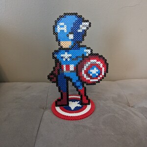 May include: A pixel art figurine of Captain America standing on a red, white, and blue circular base. The figurine is made of blue, red, white, and black beads. Captain America is holding his shield in his right hand.