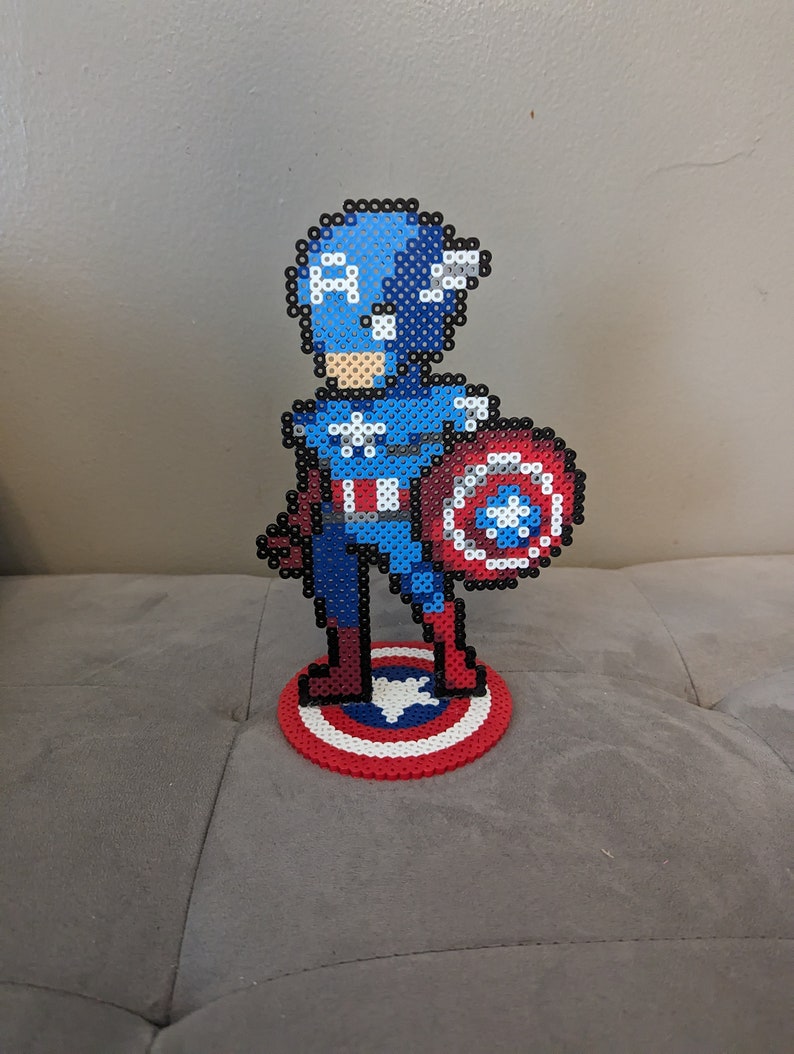 May include: A pixel art figurine of Captain America standing on a red, white, and blue circular base. The figurine is made of blue, red, white, and black beads. Captain America is holding his shield in his right hand.
