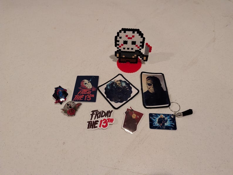 May include: A collection of Friday the 13th themed items including a pixel art figure of a hockey-masked character, several stickers featuring the movie title and character imagery, a rectangular patch, and a keychain with a small black tassel.
