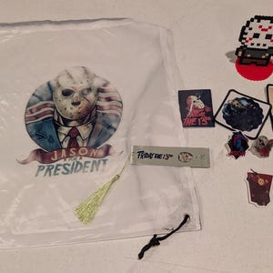 May include: A white drawstring bag featuring a graphic of a hockey-masked character in a suit with the text JASON FOR PRESIDENT, accompanied by a pixel art figure, several stickers, a metal bookmark with FRIDAY THE 13TH, and a keychain.