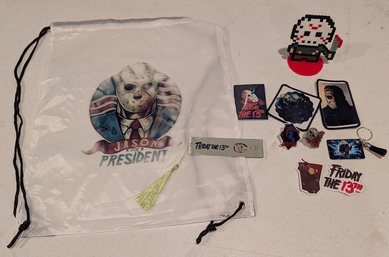 May include: A white drawstring bag featuring a graphic of a hockey-masked character in a suit with the text JASON FOR PRESIDENT, accompanied by a pixel art figure, several stickers, a metal bookmark with FRIDAY THE 13TH, and a keychain.