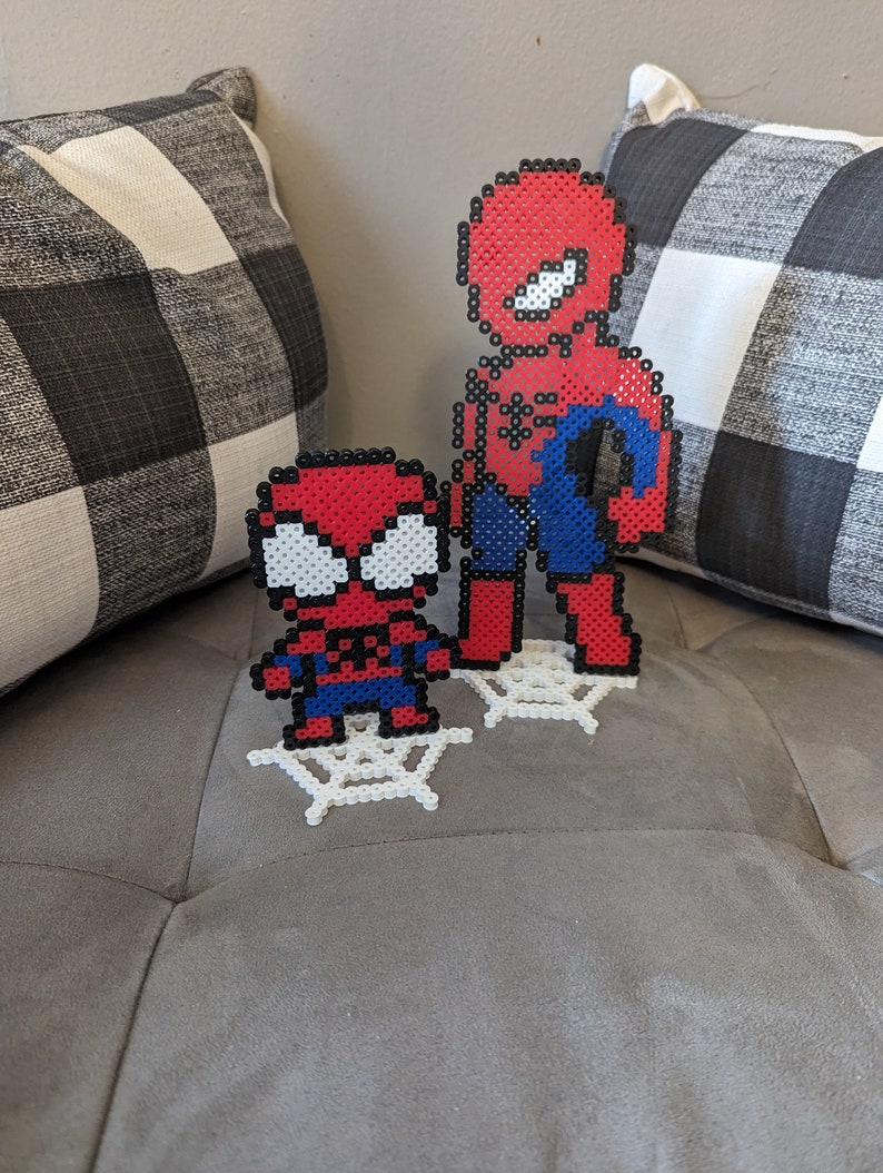 Puede incluir: Dos figuras de Spider-Man en pixel art rojo y azul de pie sobre telara&ntilde;as de pixel art blancas. El Spider-Man m&aacute;s grande est&aacute; de pie detr&aacute;s del Spider-Man m&aacute;s peque&ntilde;o.