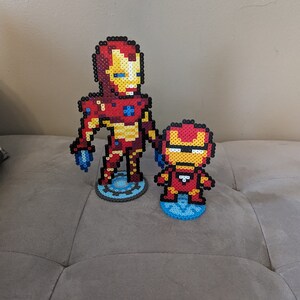 May include: Two Iron Man figures made of red, yellow, and blue perler beads. The larger figure is standing on a blue and gray circular base, while the smaller figure is standing on a blue circular base.