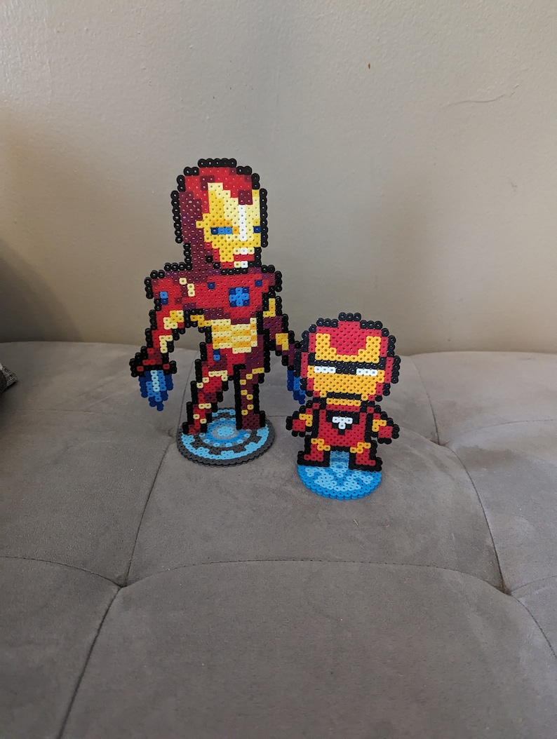 May include: Two Iron Man figures made of red, yellow, and blue perler beads. The larger figure is standing on a blue and gray circular base, while the smaller figure is standing on a blue circular base.