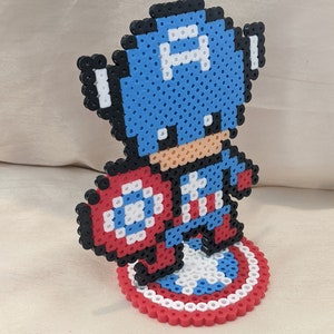 May include: A blue and red pixel art figurine of Captain America holding his shield. The figurine is standing on a circular base with a red, white, and blue design.