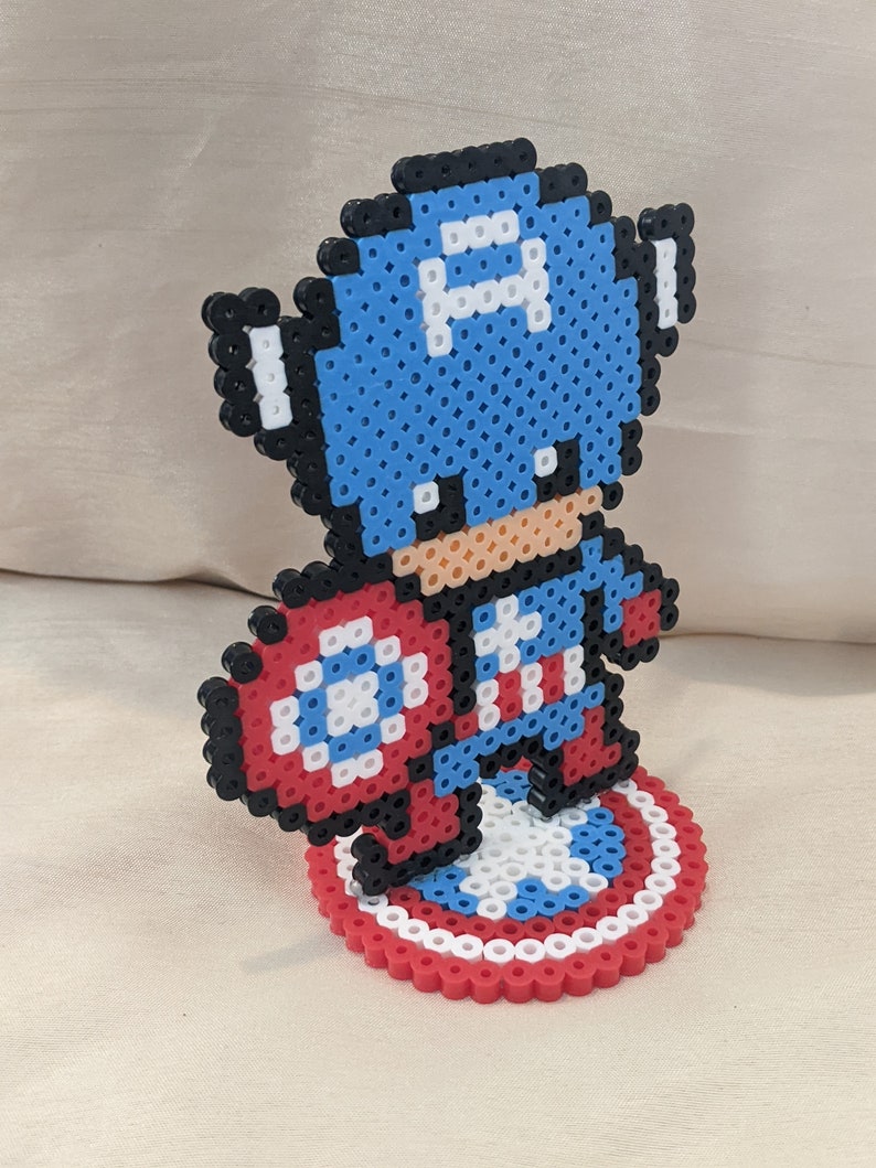 May include: A blue and red pixel art figurine of Captain America holding his shield. The figurine is standing on a circular base with a red, white, and blue design.