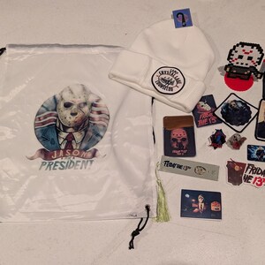 May include: A white drawstring bag featuring a Jason for President graphic, a white beanie with a Camp Crystal Lake Counselor patch, and various Friday the 13th themed items including stickers, pins, a bookmark with a tassel, and a pixel art figure.