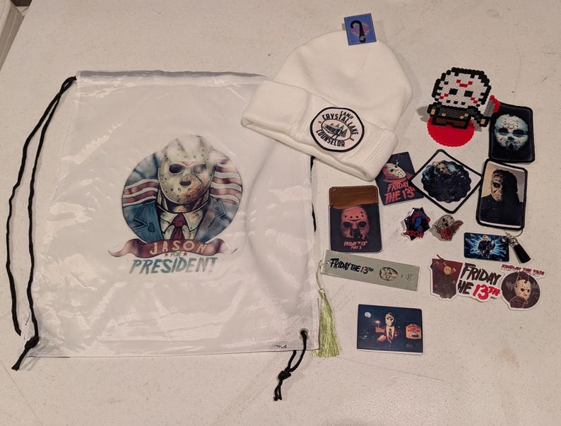 May include: A white drawstring bag featuring a Jason for President graphic, a white beanie with a Camp Crystal Lake Counselor patch, and various Friday the 13th themed items including stickers, pins, a bookmark with a tassel, and a pixel art figure.