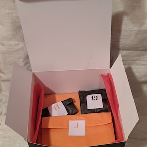 May include: A black box with a white lid is open, revealing three orange and black envelopes. The envelopes are labeled with the numbers "3", "11", and "12".