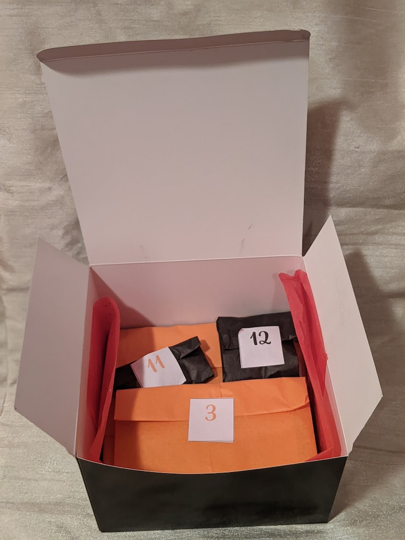 May include: A black box with a white lid is open, revealing three orange and black envelopes. The envelopes are labeled with the numbers "3", "11", and "12".