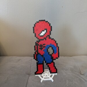 Puede incluir: Una figura de pixel art roja y azul de Spider-Man de pie sobre una telara&ntilde;a de pixel art blanca.