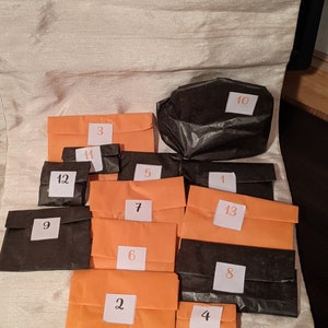 May include: Thirteen small orange and black paper bags with numbers 1 through 13 written on them. The bags are arranged in a semi-circle on a white surface.