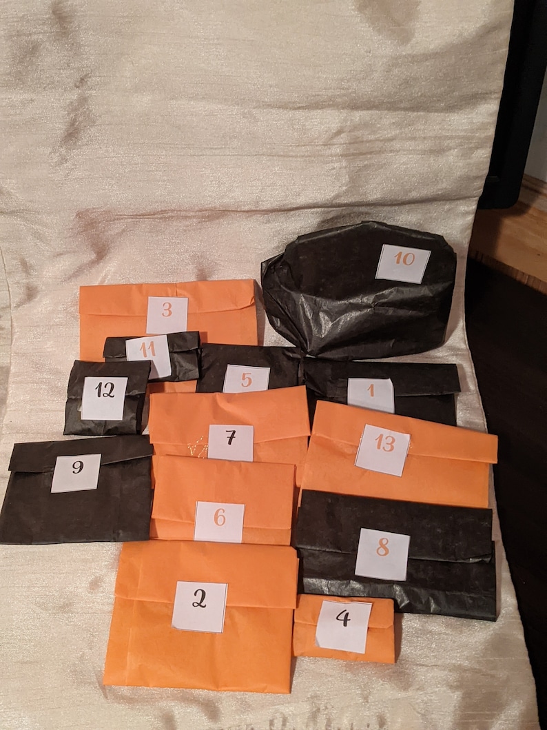 May include: Thirteen small orange and black paper bags with numbers 1 through 13 written on them. The bags are arranged in a semi-circle on a white surface.
