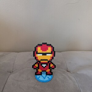 May include: A red, yellow, and black pixel art figure of Iron Man standing on a blue pixel art base.