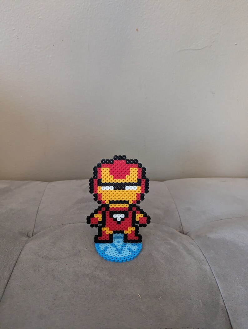 May include: A red, yellow, and black pixel art figure of Iron Man standing on a blue pixel art base.