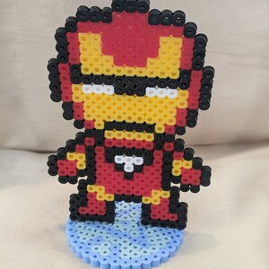 May include: A red, yellow, and black pixel art figurine of Iron Man standing on a blue pixel art base.