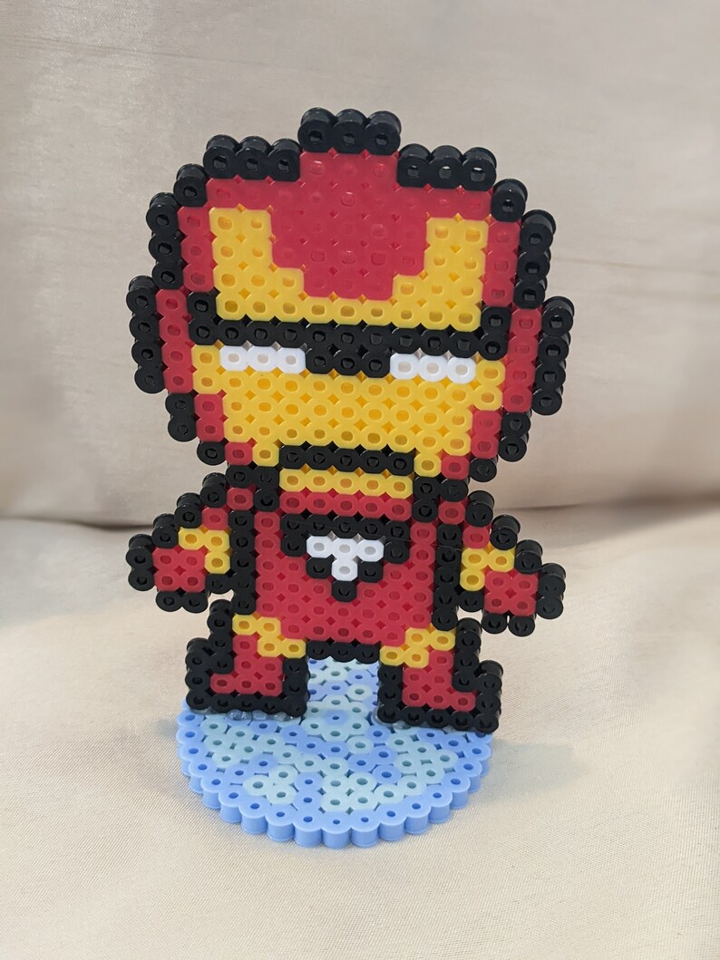 May include: A red, yellow, and black pixel art figurine of Iron Man standing on a blue pixel art base.