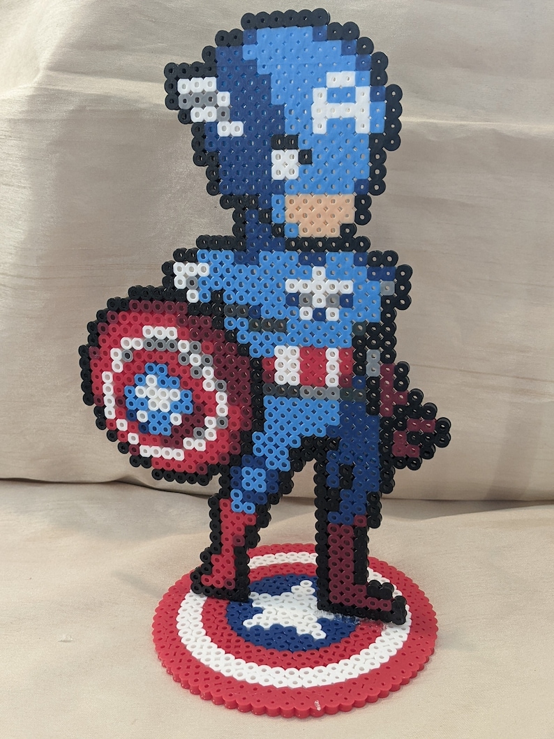 May include: A pixel art figurine of Captain America, standing with his shield in his left hand. The figurine is made of blue, red, white, and black beads. The figurine is standing on a red, white, and blue base.