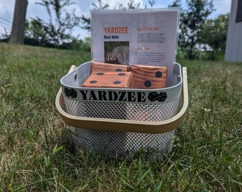 May include: A white metal basket with a wooden handle contains wooden dice with black dots. The basket is sitting on green grass. A rule book for the game Yardzee is visible in the background.