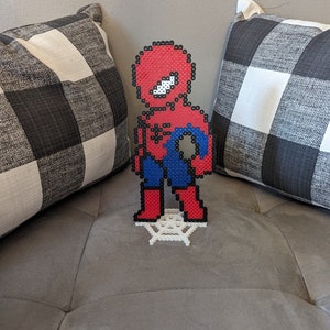Puede incluir: Una figura de pixel art de Spider-Man de color rojo y azul de pie sobre una base de telara&ntilde;a blanca. La figura est&aacute; hecha de peque&ntilde;as cuentas de pl&aacute;stico.