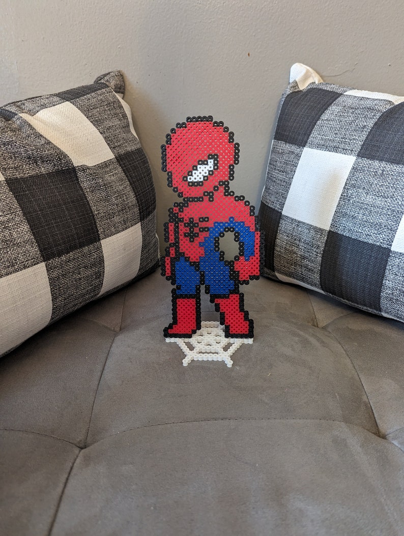 Puede incluir: Una figura de pixel art de Spider-Man de color rojo y azul de pie sobre una base de telara&ntilde;a blanca. La figura est&aacute; hecha de peque&ntilde;as cuentas de pl&aacute;stico.