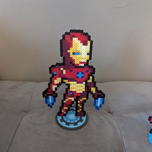 May include: A red, yellow, and blue pixel art figurine of Iron Man standing on a blue and gray pixel art base. The figurine is made of small plastic beads.
