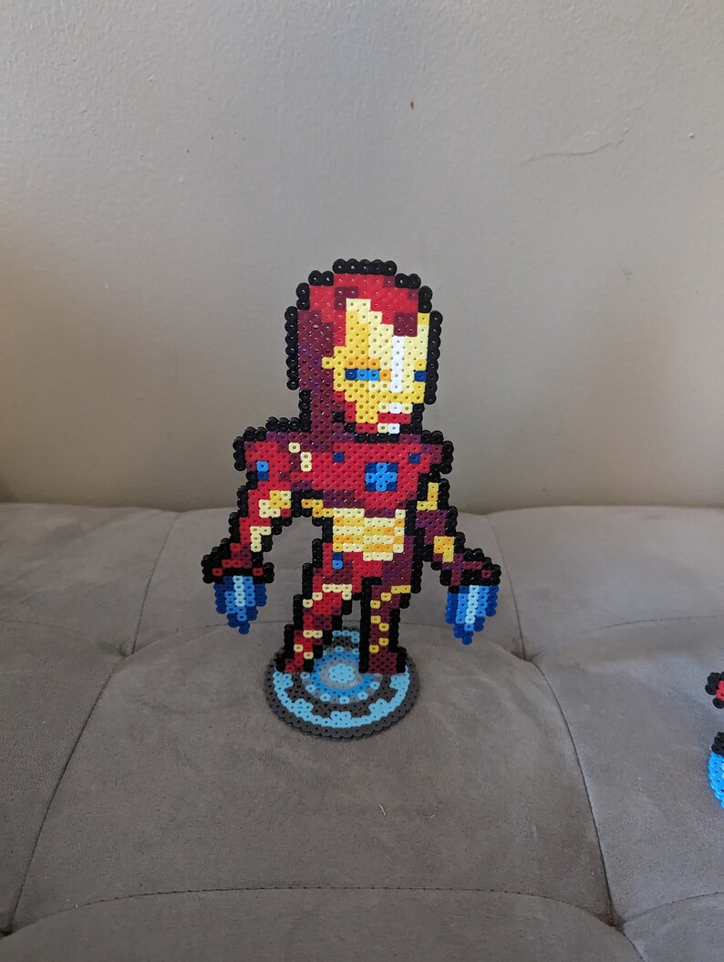 May include: A red, yellow, and blue pixel art figurine of Iron Man standing on a blue and gray pixel art base. The figurine is made of small plastic beads.