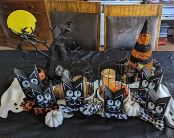 May include: A Halloween-themed table setting with black cat cutouts, pumpkins, and a witch's hat. The centerpiece includes a black tree with a yellow moon, bats, and an owl. Decorative elements include candles in wire cages and a white ghost-shaped table runner.