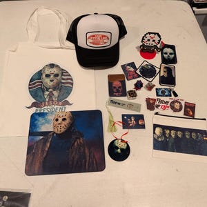 May include: A collection of Friday the 13th themed merchandise including a white tote bag with a Jason for President graphic, a black and white Camp Crystal Lake trucker hat, a mousepad, various stickers, patches, a bookmark, and a zipper pouch.