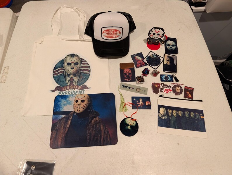 May include: A collection of Friday the 13th themed merchandise including a white tote bag with a Jason for President graphic, a black and white Camp Crystal Lake trucker hat, a mousepad, various stickers, patches, a bookmark, and a zipper pouch.