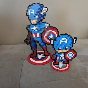 May include: Two Captain America figures made of blue, red, and white perler beads. The larger figure is standing with a shield, while the smaller figure is holding a shield and standing on a red, white, and blue circle.