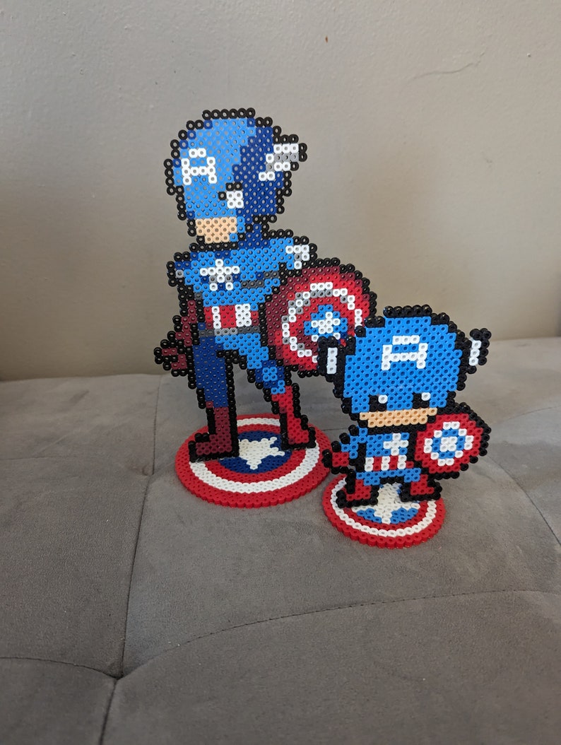 May include: Two Captain America figures made of blue, red, and white perler beads. The larger figure is standing with a shield, while the smaller figure is holding a shield and standing on a red, white, and blue circle.