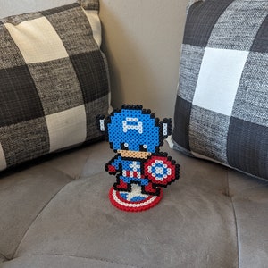 May include: A blue and red pixel art figure of Captain America standing on a red, white, and blue pixel art shield. The figure is holding a red, white, and blue pixel art shield.