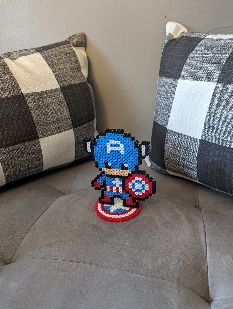May include: A blue and red pixel art figure of Captain America standing on a red, white, and blue pixel art shield. The figure is holding a red, white, and blue pixel art shield.