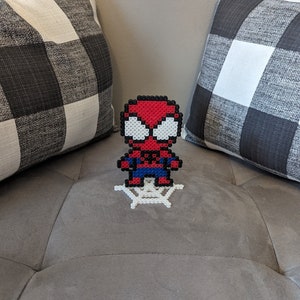 Puede incluir: Una figura de Spiderman en pixel art rojo y azul de pie sobre una telara&ntilde;a de pixel art blanca. La figura est&aacute; en un sof&aacute; gris con almohadas a cuadros blanco y negro.