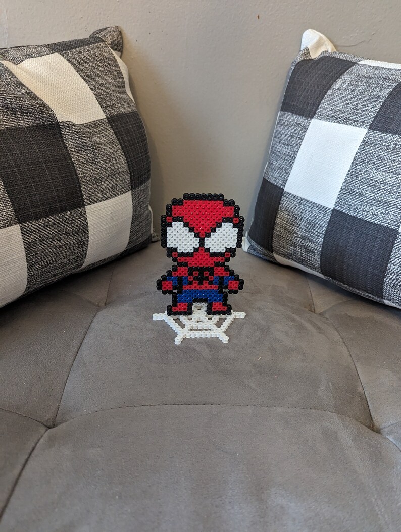 Puede incluir: Una figura de Spiderman en pixel art rojo y azul de pie sobre una telara&ntilde;a de pixel art blanca. La figura est&aacute; en un sof&aacute; gris con almohadas a cuadros blanco y negro.