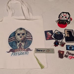 May include: A white tote bag featuring a graphic of a hockey mask-wearing character with the text JASON FOR PRESIDENT, accompanied by a pixel art figure, several rectangular stickers, a metal bookmark with a tassel, and a keychain with a charm.