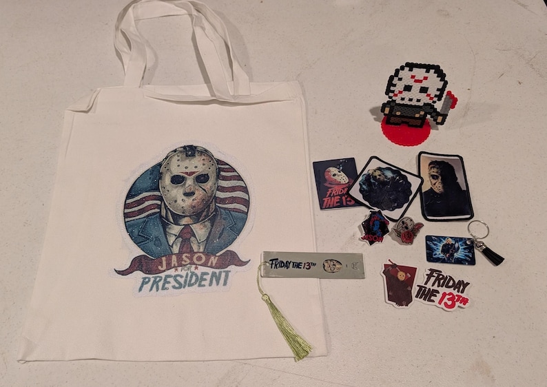 May include: A white tote bag featuring a graphic of a hockey mask-wearing character with the text JASON FOR PRESIDENT, accompanied by a pixel art figure, several rectangular stickers, a metal bookmark with a tassel, and a keychain with a charm.