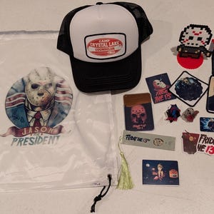 May include: A white drawstring bag with a Jason Voorhees graphic and text reading JASON FOR PRESIDENT, a black and white trucker hat with a CAMP CRYSTAL LAKE COUNSELOR patch, and various Friday the 13th themed stickers, pins, and a bookmark.