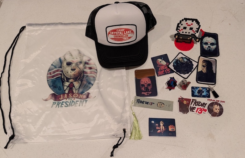 May include: A white drawstring bag with a Jason Voorhees graphic and text reading JASON FOR PRESIDENT, a black and white trucker hat with a CAMP CRYSTAL LAKE COUNSELOR patch, and various Friday the 13th themed stickers, pins, and a bookmark.