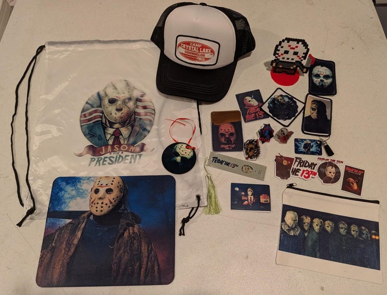 May include: A collection of Friday the 13th themed merchandise including a white drawstring bag with a Jason for President graphic, a black and white Camp Crystal Lake trucker hat, a mousepad, a zippered pouch, stickers, and various small accessories.