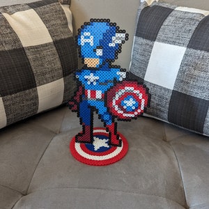 May include: A pixel art figurine of Captain America holding his shield. The figurine is made of blue, red, white, and black beads. The figurine is standing on a red, white, and blue beaded base.