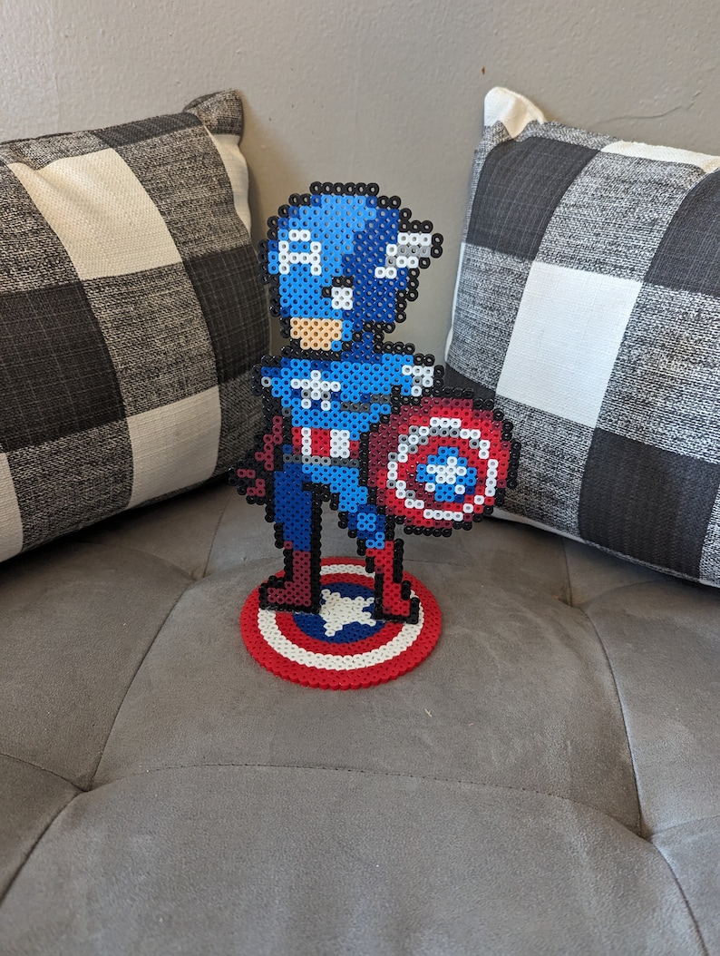 May include: A pixel art figurine of Captain America holding his shield. The figurine is made of blue, red, white, and black beads. The figurine is standing on a red, white, and blue beaded base.