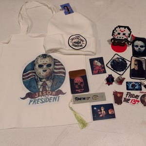 May include: A white tote bag featuring a Jason for President graphic, a white beanie with a Camp Crystal Lake Counselor patch, and various Friday the 13th themed items including stickers, pins, a bookmark with a tassel, and a pixel art figure.