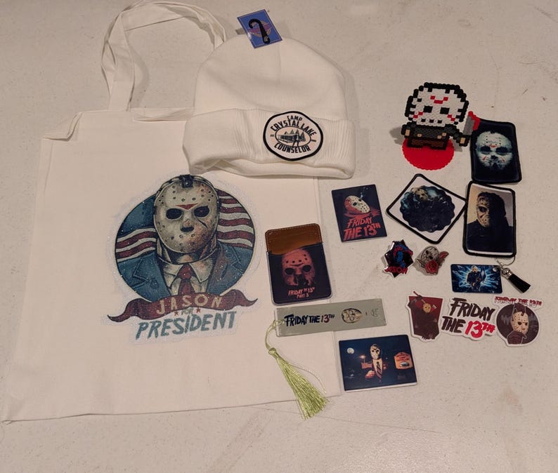 May include: A white tote bag featuring a Jason for President graphic, a white beanie with a Camp Crystal Lake Counselor patch, and various Friday the 13th themed items including stickers, pins, a bookmark with a tassel, and a pixel art figure.