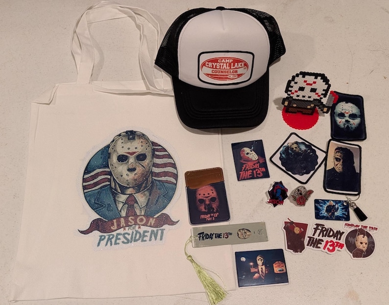 May include: A collection of horror-themed merchandise including a white tote bag with a Jason Voorhees graphic, a black and white Camp Crystal Lake trucker hat, various stickers, patches, a bookmark with a tassel, and a pixel art figure.